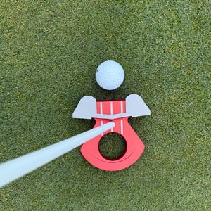 May include: A golf putting aid with a red base and a white putter. A white golf ball sits above the red base. The putting aid has a circular opening and two gray wings. The putting aid is on a green surface.