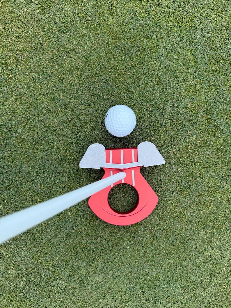 May include: A golf putting aid with a red base and a white putter. A white golf ball sits above the red base. The putting aid has a circular opening and two gray wings. The putting aid is on a green surface.