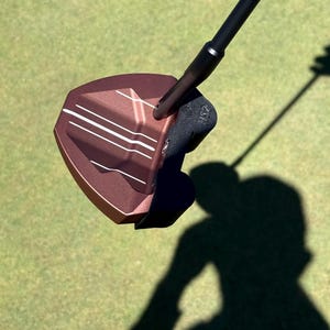 Puede incluir: Un putter de golf de color bronce con un eje y empu&ntilde;adura negros. La cabeza del putter tiene l&iacute;neas de alineaci&oacute;n blancas y una superficie texturizada. La imagen est&aacute; tomada en un campo de golf verde, con la sombra del putter visible.