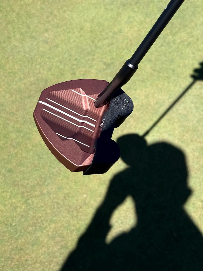 Puede incluir: Un putter de golf de color bronce con un eje y empu&ntilde;adura negros. La cabeza del putter tiene l&iacute;neas de alineaci&oacute;n blancas y una superficie texturizada. La imagen est&aacute; tomada en un campo de golf verde, con la sombra del putter visible.