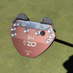 Puede incluir: Primer plano de un putter de golf con una cabeza de color cobre. El putter tiene el texto "OZ! HS" y "LAB GOLF" impreso. El putter est&aacute; sobre una superficie verde.