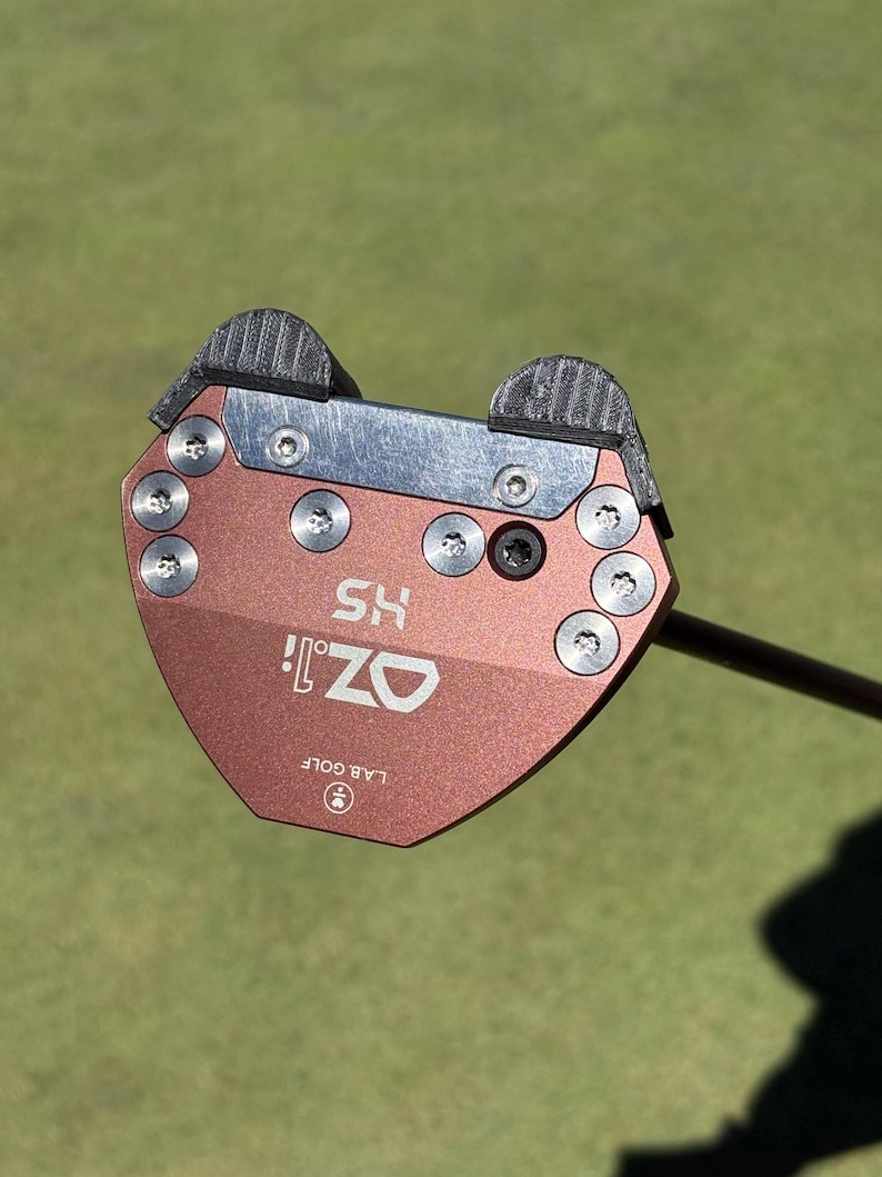 Puede incluir: Primer plano de un putter de golf con una cabeza de color cobre. El putter tiene el texto "OZ! HS" y "LAB GOLF" impreso. El putter est&aacute; sobre una superficie verde.