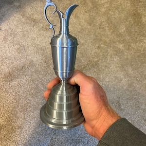 Puede incluir: Un trofeo de metal plateado con un asa decorativa y una base escalonada. El trofeo tiene forma de jarra.