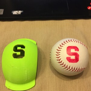 Puede incluir: Una gorra de béisbol verde con una "S" blanca en la parte delantera, junto a una pelota de béisbol blanca con una "S" roja.