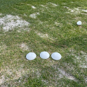 May include: Three white golf tee markers on a green grass surface with a white golf ball in the background.