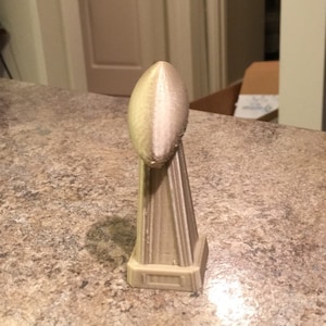 Könnte beinhalten: Eine kleine, beigefarbene Plastik-Replik des Vince Lombardi Trophy, einer fußballförmigen Trophäe, die dem Gewinner des Super Bowls verliehen wird.