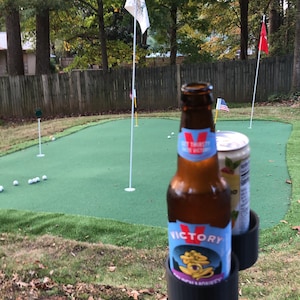 May include: A green putting green with golf balls and two flags. A black and orange golf ball holder with a bottle of Victory beer and a can of soda.