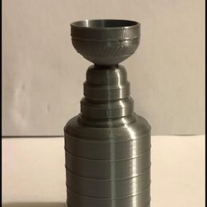 May include: A silver-colored, 3D-printed trophy cup featuring a wide, circular base, a multi-tiered cylindrical body that tapers toward the top, and a bowl-shaped top section with a textured rim. The object has a smooth, metallic finish.