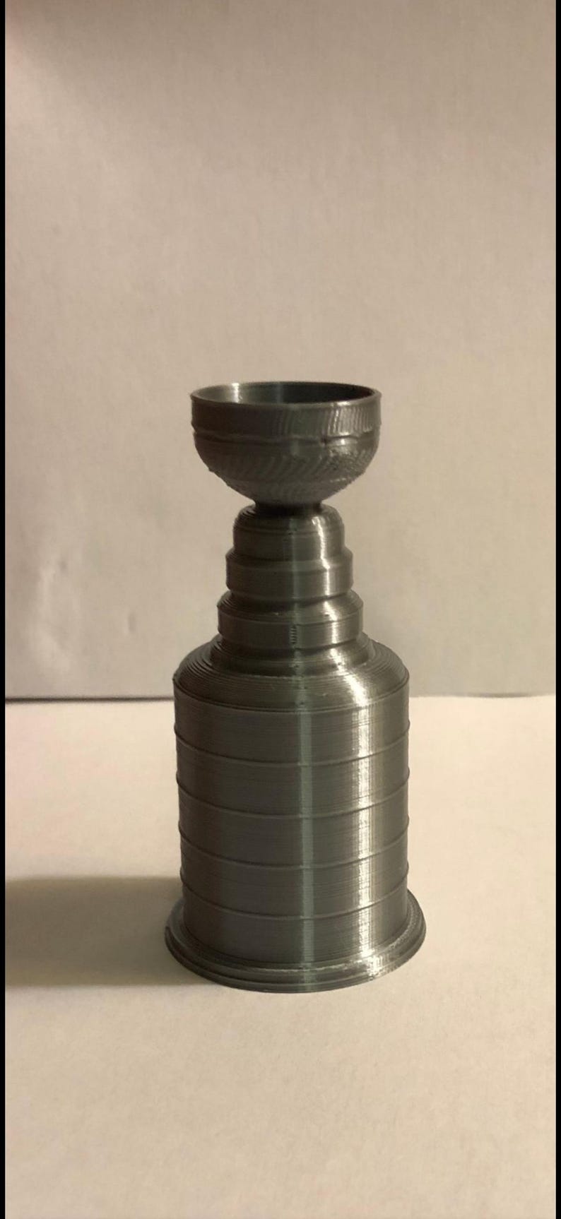 May include: A silver-colored, 3D-printed trophy cup featuring a wide, circular base, a multi-tiered cylindrical body that tapers toward the top, and a bowl-shaped top section with a textured rim. The object has a smooth, metallic finish.