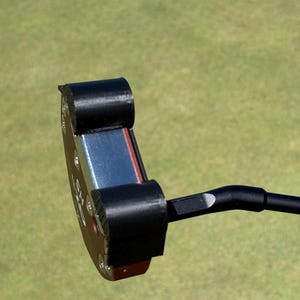 以下が含まれることがあります： 赤と青の面を持つ黒いゴルフパター。 パターヘッドは丸みを帯びた形状で、黒いグリップとシャフトが付いています。 背景はぼやけた緑のゴルフコースです。