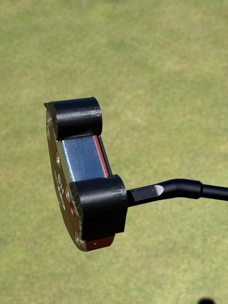 Puede incluir: Un putter de golf negro con una cara roja y azul. La cabeza del putter tiene una forma redondeada con un agarre y un eje negros. El fondo es un campo de golf verde borroso.