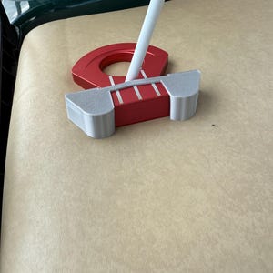 Puede incluir: Un putter de golf rojo y plateado con un eje blanco. El putter tiene una cabeza rectangular con tres líneas blancas y una pieza roja circular. El putter está sobre una superficie beige.