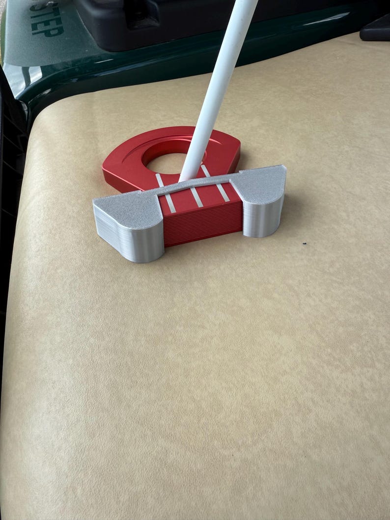 May include: A red and silver golf putter with a white shaft. The putter has a rectangular head with three white lines and a circular red piece. The putter is resting on a tan surface.