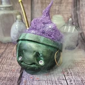 Puede incluir: Un caldero artesanal caprichoso con una cara sonriente y pendientes de aro dorados. Está lleno de espuma morada texturizada y una varita dorada. Botellas decorativas y una superficie de madera completan la escena.