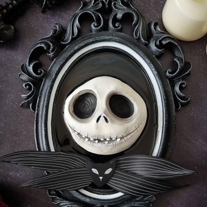 Puede incluir: Decoración de pared de Halloween en blanco y negro, con una cara de calavera sonriente dentro de un marco negro decorativo. Debajo, una pajarita negra con una silueta de gato. Diseño de estética gótica y espeluznante.