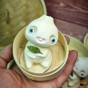 Puede incluir: Una figurita de cer&aacute;mica blanca de una criatura con grandes ojos azules y una sonrisa de dientes, sosteniendo una hoja verde. La figurita est&aacute; sentada dentro de una peque&ntilde;a cesta de vapor redonda de madera.