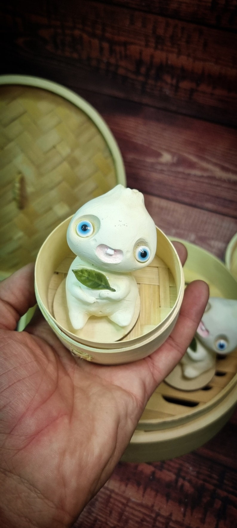 Puede incluir: Una figurita de cer&aacute;mica blanca de una criatura con grandes ojos azules y una sonrisa de dientes, sosteniendo una hoja verde. La figurita est&aacute; sentada dentro de una peque&ntilde;a cesta de vapor redonda de madera.