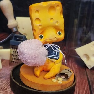 Puede incluir: Una figurita amarilla con forma de queso que sostiene una bola de algod&oacute;n de az&uacute;car rosa, con una camisa de marinero azul y blanca con un dise&ntilde;o de ancla. La figurita est&aacute; sentada sobre una base amarilla con una bolsa de papel marr&oacute;n y monedas de oro.