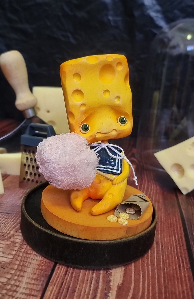 Puede incluir: Una figurita amarilla con forma de queso que sostiene una bola de algod&oacute;n de az&uacute;car rosa, con una camisa de marinero azul y blanca con un dise&ntilde;o de ancla. La figurita est&aacute; sentada sobre una base amarilla con una bolsa de papel marr&oacute;n y monedas de oro.