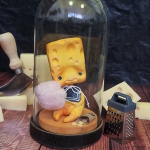 Puede incluir: Una figurita amarilla con forma de queso con un traje de marinero se sienta dentro de una c&uacute;pula de vidrio sobre una base de madera. La figurita sostiene una peque&ntilde;a nube blanca. Un rallador de plata se encuentra sobre la superficie de madera junto a la c&uacute;pula.