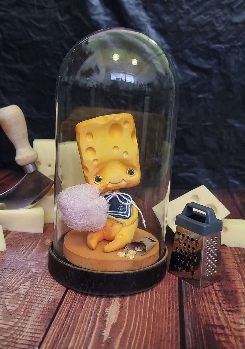Puede incluir: Una figurita amarilla con forma de queso con un traje de marinero se sienta dentro de una c&uacute;pula de vidrio sobre una base de madera. La figurita sostiene una peque&ntilde;a nube blanca. Un rallador de plata se encuentra sobre la superficie de madera junto a la c&uacute;pula.