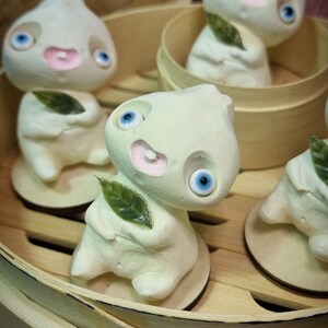 Puede incluir: Cuatro peque&ntilde;as figuritas de cer&aacute;mica blanca con ojos azules y bocas rosas est&aacute;n dispuestas en una vaporera de bamb&uacute;. Cada figurita sostiene una peque&ntilde;a hoja verde.