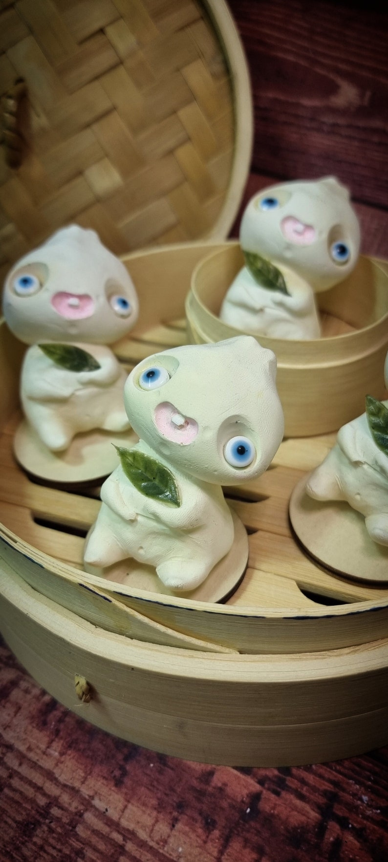 Puede incluir: Cuatro peque&ntilde;as figuritas de cer&aacute;mica blanca con ojos azules y bocas rosas est&aacute;n dispuestas en una vaporera de bamb&uacute;. Cada figurita sostiene una peque&ntilde;a hoja verde.