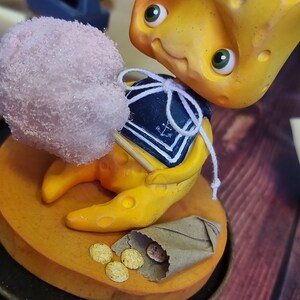 Puede incluir: Una figurita amarilla con forma de queso que lleva un traje de marinero azul y blanco sostiene un algod&oacute;n de az&uacute;car rosa en un palo. La figurita est&aacute; sentada sobre una base marr&oacute;n con una bolsa de papel marr&oacute;n y monedas de oro.