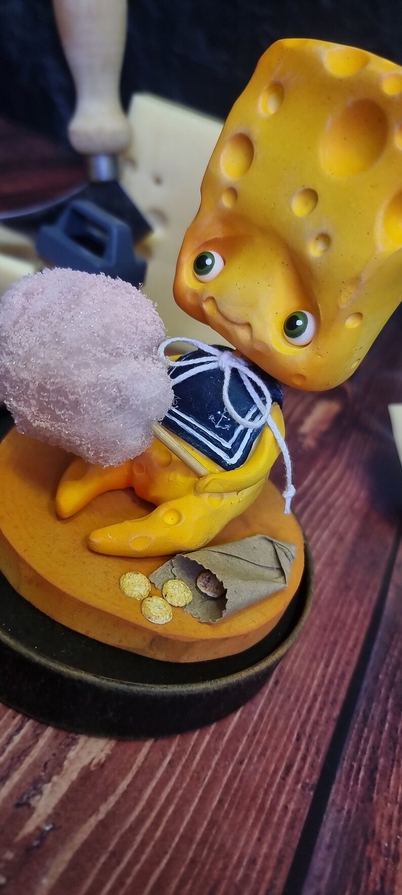 Puede incluir: Una figurita amarilla con forma de queso que lleva un traje de marinero azul y blanco sostiene un algod&oacute;n de az&uacute;car rosa en un palo. La figurita est&aacute; sentada sobre una base marr&oacute;n con una bolsa de papel marr&oacute;n y monedas de oro.