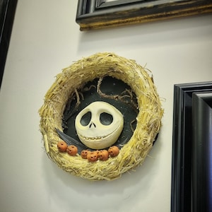 Könnte beinhalten: Ein Strohkranz mit einem Jack Skellington Gesicht und orangefarbenen Kürbissen. Der Kranz hängt an einer weißen Wand.