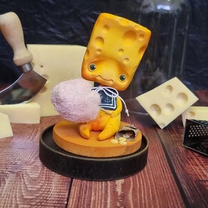 Puede incluir: Una figurita amarilla con forma de queso que sostiene un objeto rosa parecido a algod&oacute;n de az&uacute;car. La figurita lleva una camisa de marinero azul y blanca con un dise&ntilde;o de ancla. La figurita est&aacute; sentada sobre una base de madera con una peque&ntilde;a bolsa de monedas de oro.