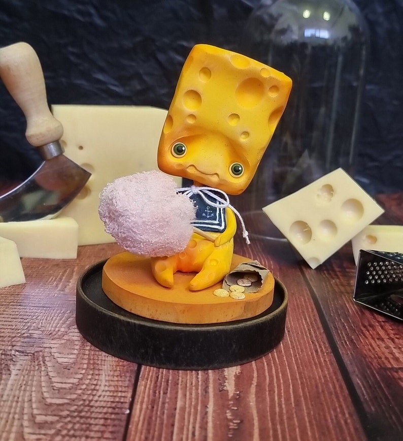Puede incluir: Una figurita amarilla con forma de queso que sostiene un objeto rosa parecido a algod&oacute;n de az&uacute;car. La figurita lleva una camisa de marinero azul y blanca con un dise&ntilde;o de ancla. La figurita est&aacute; sentada sobre una base de madera con una peque&ntilde;a bolsa de monedas de oro.