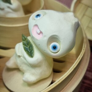 Puede incluir: Dos figuritas de cer&aacute;mica blanca con ojos azules y bocas rosas, que se asemejan a alb&oacute;ndigas, se muestran en una cesta de vapor de madera. Las figuritas sostienen hojas verdes.