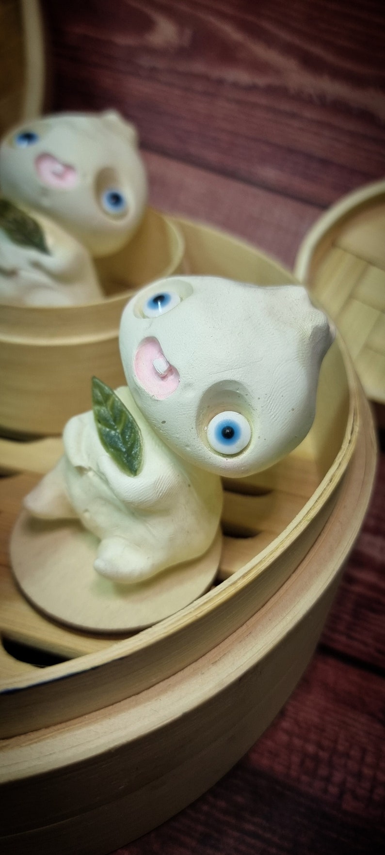 Puede incluir: Dos figuritas de cer&aacute;mica blanca con ojos azules y bocas rosas, que se asemejan a alb&oacute;ndigas, se muestran en una cesta de vapor de madera. Las figuritas sostienen hojas verdes.