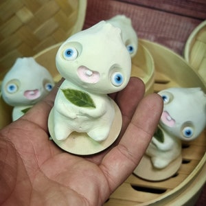 Puede incluir: Cuatro figuritas de cer&aacute;mica blanca con grandes ojos azules y mejillas rosadas. Las figuritas tienen forma de alb&oacute;ndigas y sostienen hojas verdes. Las figuritas est&aacute;n sentadas en una vaporera de bamb&uacute;.