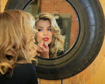 May include: A woman with long blonde hair applies red lipstick in front of a mirror. The mirror is in a tire.