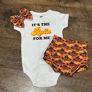 May include: A white baby onesie with the text "It's the Rolls for Me" in yellow and orange lettering. The onesie is paired with orange and brown shorts featuring a repeated pattern of "Texas" text and a hot dog graphic.
