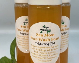 May include: Three bottles of amber-colored liquid face wash foam with white pump tops. The bottles are labeled "Sea Moss Face Wash Foam Brightening Effect" with a list of organic ingredients. The bottles are sitting on a brown surface with green leaves.