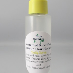 K&ouml;nnte beinhalten: Eine durchsichtige Kunststoffspr&uuml;hflasche mit gelbem Spr&uuml;hkopf, die Brina Organics Fermented Rice Water & Biotin Hair Hydrate enth&auml;lt. Das Etikett listet Inhaltsstoffe wie fermentierten Reis, Biotin und Bio-Ylang-Ylang auf.