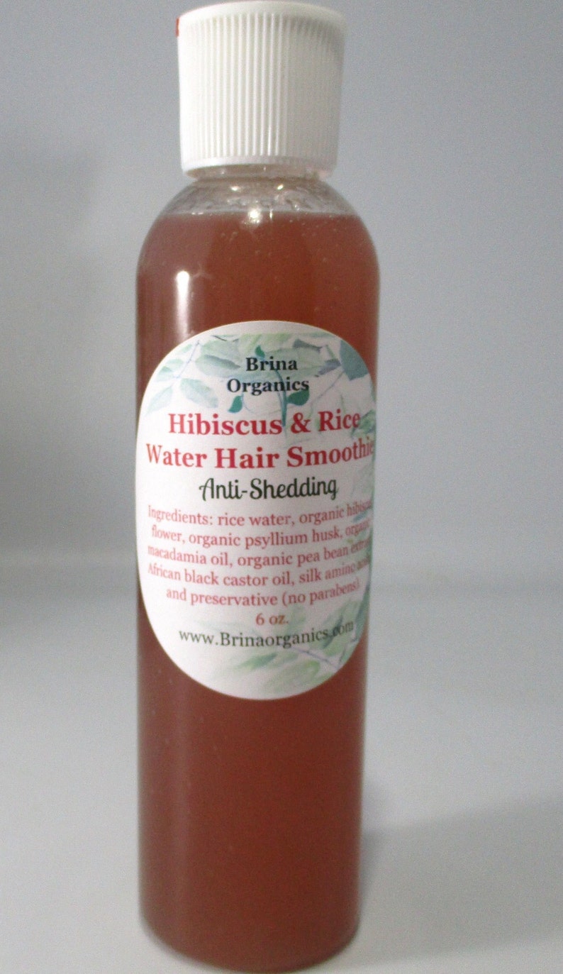 May include: A clear plastic bottle containing a reddish-brown liquid hair product with a white cap. The label reads Brina Organics Hibiscus & Rice Water Hair Smoothie Anti-Shedding, listing ingredients and a 6 oz. volume, with a green leaf design.