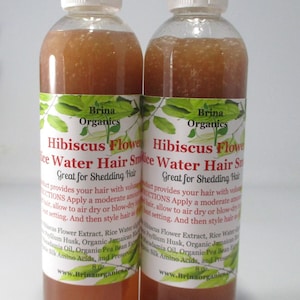 May include: Two clear plastic bottles containing a brownish liquid hair product. The labels feature green leaf illustrations and text reading Brina Organics Hibiscus Flower Rice Water Hair Smoothie, Great for Shedding Hair, and 8 oz.