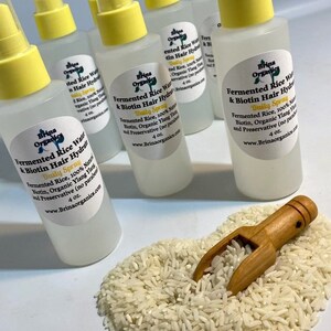 K&ouml;nnte beinhalten: Mehrere durchsichtige Kunststoff-Spr&uuml;hflaschen mit gelben Verschl&uuml;ssen, die jeweils 118 ml Fermented Rice Water & Biotin Hair Hydration Daily Spray enthalten. Die Etiketten f&uuml;hren fermentierten Reis, Biotin und Ylang-Ylang auf. Eine kleine Holzschaufel liegt in einem Haufen wei&szlig;er Reisk&ouml;rner.