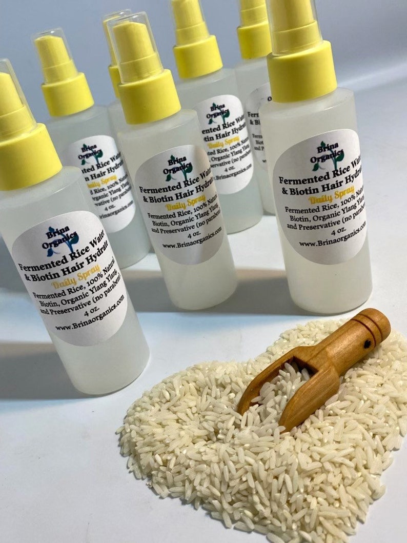K&ouml;nnte beinhalten: Mehrere durchsichtige Kunststoff-Spr&uuml;hflaschen mit gelben Verschl&uuml;ssen, die jeweils 118 ml Fermented Rice Water & Biotin Hair Hydration Daily Spray enthalten. Die Etiketten f&uuml;hren fermentierten Reis, Biotin und Ylang-Ylang auf. Eine kleine Holzschaufel liegt in einem Haufen wei&szlig;er Reisk&ouml;rner.