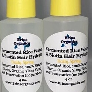 K&ouml;nnte beinhalten: Zwei durchsichtige Kunststoff-Spr&uuml;hflaschen mit gelben Kappen, die jeweils 118 ml Brina Organics Fermented Rice Water & Biotin Hair Hydrate Daily Spray enthalten. Die wei&szlig;en ovalen Etiketten f&uuml;hren Inhaltsstoffe wie Reiswasser auf.