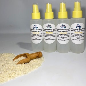 K&ouml;nnte beinhalten: Vier durchsichtige Kunststoffspr&uuml;hflaschen mit gelben Kappen, jeweils beschriftet mit Brina Organics Fermented Rice Water & Biotin Hair Hydrate Daily Spray 118 ml. Eine kleine Holzschaufel liegt in einem Haufen wei&szlig;er Reisk&ouml;rner.