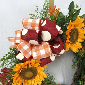 Pode incluir: Uma coroa de outono com um grande la&ccedil;o feito de fita xadrez laranja e branca e fita bord&ocirc; com bolinhas brancas. A coroa &eacute; decorada com girass&oacute;is e folhagem.