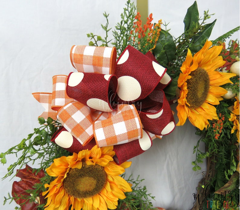 Pode incluir: Uma coroa de outono com um grande la&ccedil;o feito de fita xadrez laranja e branca e fita bord&ocirc; com bolinhas brancas. A coroa &eacute; decorada com girass&oacute;is e folhagem.