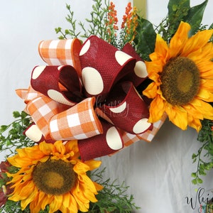 Pode incluir: Uma coroa de outono com um grande la&ccedil;o vermelho com bolinhas brancas e um la&ccedil;o menor de xadrez laranja e branco. A coroa &eacute; decorada com girass&oacute;is e vegeta&ccedil;&atilde;o.