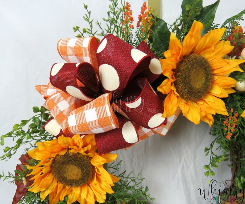 Pode incluir: Uma coroa de outono com um grande la&ccedil;o vermelho com bolinhas brancas e um la&ccedil;o menor de xadrez laranja e branco. A coroa &eacute; decorada com girass&oacute;is e vegeta&ccedil;&atilde;o.