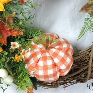 Pode incluir: Uma coroa de outono com base de videira marrom, decorada com girass&oacute;is, folhas verdes, flores brancas e uma pequena ab&oacute;bora xadrez laranja e branca.
